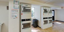 This shared laundry facility is designed with practicality and efficiency in mind, providing residents with a well-equipped space for their laundry needs. The room features a row of commercial-grade front-loading washing machines lining one wall, complemented by several stacked dryer units by Hercules positioned throughout the space. The machines are numbered for easy identification, and usage instructions are clearly posted on the walls for resident reference.

The facility is finished with durable tile flooring throughout, providing an easy-to-clean surface that withstands heavy foot traffic. White walls and fluorescent overhead lighting keep the space bright and functional. Natural light filters in through windows along one wall, offering views of surrounding greenery and adding a welcoming element to the utilitarian environment.

A small seating area with two blue chairs is available for residents waiting on their laundry. The entry area includes a payment kiosk, a fire extinguisher, and an exit sign, reflecting the facility's attention to safety and convenience. A folding table provides a practical surface for handling laundry. The overall layout is open and uncluttered, ensuring residents can navigate the space with ease.