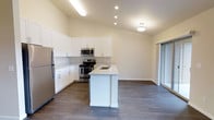 This single-floor space showcases a thoughtfully designed open-concept layout that seamlessly connects the kitchen and living areas. The vaulted ceiling rises to an impressive peak, lending the space a sense of openness and airiness that is further enhanced by recessed lighting and a central flush-mount fixture.

The kitchen is a standout feature, outfitted with crisp white shaker-style cabinetry that extends to the upper walls for ample storage. A full suite of stainless steel appliances—including a refrigerator, electric range, and microwave—provides a sleek, modern aesthetic. The central kitchen island anchors the space, featuring a light-toned countertop with an integrated sink, offering both additional prep space and a natural gathering point.

Underfoot, rich dark hardwood-style flooring runs throughout, adding warmth and contrast to the bright, neutral wall tones. A large sliding glass door adjacent to the kitchen and living area allows natural light to flow in and provides convenient access to the outdoors.

The overall ambiance of the space is clean, contemporary, and move-in ready, with a cohesive design language that balances functionality with modern style. The open floor plan and elevated ceiling make the space feel expansive and welcoming.