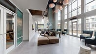 Located at 425 Hillsborough St in Chapel Hill, NC, this contemporary amenity space makes an immediate impression with its soaring double-height ceilings and expansive floor-to-ceiling windows that flood the interior with natural light. Large-format porcelain tile flooring flows throughout, lending a sleek, cohesive feel to the entire space.

The main lounge area anchors the layout, featuring a generous modular sectional sofa complemented by elegant teal wingback chairs arranged beneath a striking cluster of sculptural pendant lights. This welcoming gathering area connects seamlessly to a casual seating nook with built-in banquette seating, bar-height tables, and eclectic accent stools, all framed by curated artwork on the walls.

A dedicated game room offers a full-size billiards table set beneath towering windows that overlook an outdoor terrace, while a nearby entertainment counter with built-in seating and a mounted display screen provides a versatile social hub. A foosball table adds to the recreational options available in this zone.

Patterned glass double doors partition distinct zones while maintaining visual openness throughout. A staircase with metal railings leads to an upper level, adding architectural dimension. Multiple glass doors open directly to an outdoor patio area, reinforcing the connection between interior and exterior. The overall design balances modern sophistication with a comfortable, community-focused atmosphere.