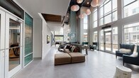 Located at 425 Hillsborough St in Chapel Hill, NC, this contemporary amenity space makes an immediate impression with its soaring double-height ceilings and expansive floor-to-ceiling windows that flood the interior with natural light. Large-format porcelain tile flooring flows throughout, lending a sleek, cohesive feel to the entire space.

The main lounge area anchors the layout, featuring a generous modular sectional sofa complemented by elegant teal wingback chairs arranged beneath a striking cluster of sculptural pendant lights. This welcoming gathering area connects seamlessly to a casual seating nook with built-in banquette seating, bar-height tables, and eclectic accent stools, all framed by curated artwork on the walls.

A dedicated game room offers a full-size billiards table set beneath towering windows that overlook an outdoor terrace, while a nearby entertainment counter with built-in seating and a mounted display screen provides a versatile social hub. A foosball table adds to the recreational options available in this zone.

Patterned glass double doors partition distinct zones while maintaining visual openness throughout. A staircase with metal railings leads to an upper level, adding architectural dimension. Multiple glass doors open directly to an outdoor patio area, reinforcing the connection between interior and exterior. The overall design balances modern sophistication with a comfortable, community-focused atmosphere.