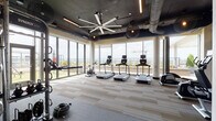 This fitness center is designed with both function and aesthetics in mind, offering a spacious, open layout that accommodates a comprehensive range of workout equipment. The space is defined by expansive floor-to-ceiling windows along one wall, flooding the interior with natural light and providing sweeping panoramic views of the surrounding skyline and landscape—an energizing backdrop for any workout.

The cardio zone features a row of treadmills, stationary bikes, and an elliptical trainer, all positioned to take full advantage of the views. A dedicated strength training area includes a multi-functional cable and rack system by Life Fitness, stocked with kettlebells, battle ropes, and various free weights. A rowing machine and additional functional fitness accessories round out the equipment offerings.

The ceiling draws immediate attention with its exposed dark industrial design, featuring visible ductwork, linear LED lighting strips, globe pendant fixtures, and a large statement ceiling fan that adds both airflow and visual impact. The flooring transitions between patterned carpet tiles in the cardio zone and rubber flooring in the strength area, providing practical surfaces suited to each activity. Glass doors open directly to an outdoor terrace, seamlessly connecting the interior space to the fresh air outside.