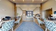 Located at 130 Canary Pl in Burlington, NC, this single-story clubhouse offers a generously sized, open-concept layout designed for both functionality and modern aesthetic appeal. The space is defined by soaring vaulted ceilings, exposed brick columns, and polished concrete floors that lend an industrial-chic character throughout.

Upon entry, glass double doors open into a welcoming reception area centered around a granite-topped credenza backed by a rich navy blue accent wall. Flanking brick columns frame the space and guide visitors into the main gathering areas. A central lounge features a curved cream sectional sofa, a bold navy geometric area rug, an ornate globe chandelier, and a fireplace — creating a warm and inviting atmosphere.

A brick-accented bar with granite countertops and barstool seating serves as a social focal point, bathed in natural light from large windows that offer outdoor views. Adjacent cabinetry and counter space provide practical utility. The space also includes a dedicated private office appointed with a statement chandelier, patterned window treatments, and ample natural light. A billiards table adds a recreational element to the open floor plan.

Throughout, navy blue accent walls, patterned upholstery, and globe pendant lighting create a cohesive, sophisticated design palette that balances comfort with refined style.