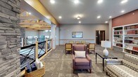 Located at 600 Gateway Place in Woodbury, Minnesota, this single-floor community space is defined by its striking architectural design and carefully curated interior spaces. The centerpiece of the layout is a grand upper-level lounge that overlooks a multi-story atrium below, connected by a staircase framed with black metal and natural wood railings. Exposed timber beam trusses span the vaulted ceiling overhead, complemented by two large circular chandelier fixtures that anchor the space with an elegant yet rustic character.

The lounge area is divided into distinct functional zones. A seating area near the staircase balcony offers plush upholstered chairs arranged around accent tables, ideal for casual conversation. A dedicated dining zone features a dark stone-topped table surrounded by wooden chairs, illuminated by recessed lighting. A separate, more intimate lounge space is highlighted by a dramatic floor-to-ceiling stacked-stone fireplace wall with a sleek linear gas insert, flanked by comfortable seating. An adjacent reading nook includes built-in bookshelves, a window bench with natural views of the outdoors, and additional seating arrangements. Patterned carpeting, muted warm tones, and layered lighting throughout contribute to an inviting, residential ambiance.