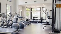 This fitness center is designed to support a comprehensive workout experience within a bright, open, and well-appointed space. The facility features soaring vaulted ceilings that lend an airy, expansive feel, complemented by an abundance of natural light streaming through large, dark-framed windows that offer views of the surrounding greenery. The flooring throughout consists of durable rubber gym flooring, practical for both cardio and strength training activities. A striking large-scale wall mural adds a bold decorative element, while full-length mirrors along one wall visually expand the space and allow users to monitor their form during workouts. A ceiling fan provides additional air circulation throughout the main exercise area. The cardio section is outfitted with multiple treadmills, elliptical trainers, and stationary bikes, including recumbent models from Precor. The strength training area includes a selection of Duo-Tek cable machines, a multi-function cable station, adjustable weight benches, and a well-stocked dumbbell rack ranging across various weights. Mounted flat-screen televisions provide entertainment during workouts. The layout is thoughtfully arranged to maximize usable floor space, offering a functional and motivating environment suitable for a wide range of fitness routines.