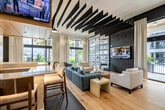 Located at 5050 Aspire Way in Naples, FL, this single-floor amenity space delivers a sophisticated social experience across a generously sized open layout. The interior is defined by striking architectural details, including dramatic black blade ceiling accents, warm light wood flooring, and a mix of tile and hardwood surfaces that delineate distinct activity zones.

At the heart of the space sits a large bar and lounge area, lined with bar-height seating and illuminated by sculptural pendant lighting. Multiple wall-mounted TVs are integrated throughout, making it ideal for gathering to watch sports or entertainment. A cozy fireplace lounge features a sleek linear electric fireplace set into a richly textured dark accent wall, flanked by plush rounded armchairs and a gold-accented coffee table.

A dedicated golf simulator room offers an immersive experience with a full projection screen, turf flooring, and club storage, accompanied by comfortable lounge seating. The billiards area showcases a full-size pool table beneath a textured white 3D accent wall and wall-mounted TV, with floor-to-ceiling windows inviting abundant natural light and views of lush Florida greenery.

Throughout the space, expansive windows and sheer drapery create a bright, airy ambiance that connects the interior to the outdoor surroundings, reinforcing the upscale, resort-inspired character of this amenity lounge.