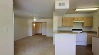 This single-floor residence features an open-concept design that seamlessly connects the kitchen and living areas, creating a spacious and functional layout. The living room is finished with wall-to-wall carpet, lending a warm and comfortable feel to the space, and is equipped with a ceiling fan to promote airflow throughout. A overhead HVAC vent is positioned in the ceiling, ensuring efficient climate control year-round. The kitchen is situated adjacent to the living area and features light wood cabinetry providing ample storage, a peninsula-style countertop that doubles as a breakfast bar or prep surface, and a white range. An under-cabinet range hood is installed above the cooking area. The countertops are a neutral tone, complementing the light, airy color palette of the unit. A hallway visible from the main living area leads to additional rooms, including what appears to be a bedroom with a closet, illuminated by overhead lighting. The walls throughout are finished in a clean, neutral white, contributing to a bright and welcoming atmosphere. The overall design prioritizes practicality and ease of living within a cohesive, open floor plan.