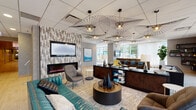 Located at 345 E Wacker Drive in the heart of Chicago, these common areas offer a thoughtfully curated environment designed for both relaxation and productivity. The central lounge serves as the focal point of the space, anchored by a teal velvet sofa and coordinating seating arrangements set atop layered area rugs. A striking textured accent wall houses a wall-mounted flat-screen TV above a sleek linear electric fireplace, creating a warm and inviting atmosphere. Overhead, dramatic large-scale starburst pendant lights draw the eye upward and lend an artistic, contemporary flair to the space. A dark wood reception-style counter provides a functional workspace with additional seating nearby. Glass-partitioned private rooms along the perimeter offer enclosed settings for focused work or small gatherings. A dedicated game and social corridor features a shuffleboard table alongside a long communal bar-height table with vibrant blue seating. A quieter reading nook, furnished with a leather sofa and bold navy accent chairs, sits beneath angular floating bookshelves and benefits from generous natural light streaming through floor-to-ceiling windows with views of the surrounding Chicago skyline and streetscape. Light wood-look flooring and textured wall finishes tie the entire space together cohesively.