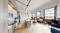 This single-floor loft-style unit offers a compelling fusion of industrial heritage and contemporary design. The open-concept kitchen and living area serves as the heart of the space, anchored by soaring ceilings with exposed whitewashed wood planking and dramatic pendant lighting suspended from a sculptural ceiling track. The kitchen is outfitted with two-tone cabinetry, a subway tile backsplash, stainless steel appliances, and a generous island with a white quartz countertop — ideal for both meal preparation and casual dining. Polished concrete-style flooring flows throughout the main area, adding to the urban character of the space. The living area is bathed in natural light from a row of tall, black-framed industrial windows with rich wood wainscoting beneath them, offering open views of the surrounding streetscape. A long, well-lit hallway with light wood flooring and a rustic whitewashed ceiling connects the main living space to the private areas, including a bedroom and a full bathroom. The bathroom features a vanity with wood cabinetry, a white countertop, and a shower-tub combination. Throughout the unit, the interplay of exposed brick-painted walls, high ceilings, and abundant natural light creates a bright, airy, and distinctly urban ambiance.