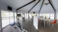 This single-story fitness center offers a well-appointed workout environment defined by its striking vaulted ceiling with bold black structural beams that draw the eye upward, adding architectural character to the space. Floor-to-ceiling windows line the walls on multiple sides, flooding the interior with abundant natural light and providing open views of the surrounding greenery, creating an airy and motivating atmosphere.

The workout area is equipped with a selection of cardio and strength training machines, including ellipticals, a cable machine, and additional cardio equipment positioned to take full advantage of the natural light and views. Dark rubberized flooring grounds the space and provides a practical, durable surface throughout the fitness area.

A ceiling fan assists with air circulation, contributing to comfort during workouts. A wall-mounted television is positioned for easy viewing from the equipment. A low partition wall separates a casual seating area furnished with a high-top table and chairs, offering a space to rest or socialize. The overall ambiance is clean, modern, and bright, making it a functional and welcoming fitness facility.