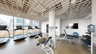 Located at 1457 North Halsted Street in Chicago's Lincoln Park neighborhood, this fitness center offers a thoughtfully designed, single-floor layout spanning a generously sized footprint. The space is defined by its striking floor-to-ceiling glass walls and large windows that flood the interior with natural light and frame expansive views of the surrounding Chicago skyline and rooftop terrace beyond.

The main workout area features a distinctive grid-pattern ceiling that adds architectural character to the open, modern aesthetic. It is equipped with a row of commercial-grade treadmills positioned directly in front of the panoramic windows, along with a multi-station cable strength machine, recumbent and upright bikes, elliptical trainers, a weight bench, and a stability ball, providing a comprehensive range of cardio and strength training options. A wall-mounted flat-screen TV is also present for entertainment during workouts.

A separate glass-enclosed room, clearly marked, offers a private area for additional exercise or stretching. The entry corridor features large-format light tile flooring and leads to double glass exterior doors that open to the outdoor rooftop deck area. Throughout the facility, the clean white walls, polished concrete floors in the gym area, and abundant natural light create a bright, motivating, and professional environment well-suited for daily fitness routines.