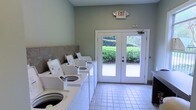 This single-floor communal laundry facility is designed with practicality and convenience in mind. The room is finished with light sage-green walls and durable white tile flooring, creating a clean and welcoming environment. A row of top-loading washing machines lines one wall, accompanied by a tile backsplash that adds a touch of polish to the functional space. Natural light fills the room through a set of white-framed French doors, which provide direct access to an outdoor area surrounded by lush greenery, offering pleasant views and easy ventilation. The doors are fitted with a standard door closer for convenience and security. An illuminated exit sign is prominently mounted above the doors, ensuring clear safety guidance. The layout is open and uncluttered, allowing for easy movement throughout the space. Overall, the facility presents a tidy, well-maintained environment that prioritizes user comfort and accessibility.