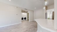 This clean, modern single-floor home is designed with a bright, neutral palette and thoughtful layout throughout. The open-concept kitchen serves as a central highlight, featuring white shaker-style cabinetry, sleek quartz countertops, a large peninsula island, stainless steel appliances including a refrigerator, range, and dishwasher, and a professional-style faucet over an undermount sink. Pendant lighting with glass fixtures adds a refined touch overhead, while light wood-look flooring flows seamlessly through the main living and kitchen areas.

The living space is generously proportioned and connects directly to a covered outdoor patio through full-width sliding glass doors, allowing abundant natural light and views of surrounding greenery. A ceiling fan with integrated lighting provides both comfort and style.

The bedrooms feature soft wall-to-wall carpeting, ceiling fans, and windows that invite natural light. One bedroom includes access to a walk-in closet fitted with built-in corner shelving, offering excellent storage. A dedicated interior laundry room houses a full-size front-loading washer and dryer, with an adjacent bathroom visible nearby. Throughout the home, clean white trim, recessed lighting, and neutral warm-toned walls create a cohesive, move-in-ready atmosphere.