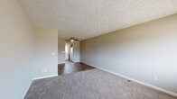 This single-floor residence offers a practical and well-organized layout across its living spaces. The main living area is generously sized, featuring neutral gray walls, soft wall-to-wall carpeting, and a textured ceiling with a ceiling fan overhead. The flooring transitions from carpet in the living area to dark hardwood-style planks in the connecting spaces, creating a clear visual distinction between zones.

The kitchen is a functional galley-style space with warm wood cabinetry, light granite-style countertops, a double stainless steel sink, and a white appliance suite. A paneled exterior door with a window allows natural light to filter in, brightening the workspace.

The bedrooms are carpeted and finished with the same cool gray palette, each featuring windows fitted with horizontal blinds that provide both privacy and natural light. The rooms offer ample wall space suited for a variety of furniture arrangements.

The bathroom is clean and efficiently designed, featuring a full-size white soaking tub and shower combination with built-in shelving, complemented by dark hardwood-style flooring and warm taupe walls. Chrome fixtures and accessories complete the space.

Overall, the residence presents a cohesive, move-in ready aesthetic with consistent neutral finishes, practical flooring choices, and a straightforward floor plan that maximizes usability throughout.
