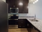 This well-appointed single-floor residence at 650 Route 23 in Wayne, New Jersey offers a thoughtfully designed layout that balances comfort and functionality. The kitchen is a standout feature, showcasing dark espresso cabinetry, granite countertops, stainless steel appliances including a refrigerator, electric range, microwave, and dishwasher, along with hardwood-style flooring and track lighting overhead. The kitchen opens via a breakfast bar into the main living area, which is finished with plush neutral carpeting and flows into additional living space featuring built-in shelving, a media niche, and a door leading to the outdoors, allowing for natural light and outdoor access. The spacious bedroom is carpeted, filled with natural light through a large window, and includes a ceiling fan and a closet. The bathroom is well-equipped with dark-toned cabinetry, granite countertops, a framed mirror, hardwood-style flooring, a tub-shower combo with white tile surround, and a private water closet. Throughout the unit, neutral wall tones and clean finishes create a bright, cohesive ambiance suited for comfortable everyday living.