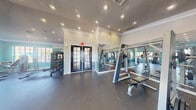 Located at 5555 North Military Trail in Boca Raton, FL, this fitness center offers a well-appointed, single-floor workout facility designed to support a comprehensive exercise routine. The space is defined by its elegant architectural details, including crown molding, a textured ceiling, and recessed lighting that creates a bright, energizing atmosphere throughout.

The fitness floor is covered in durable, dark rubber flooring suitable for a wide range of exercise activities. Large multi-pane windows line the walls, flooding the space with natural light and offering pleasant views of the surrounding Florida landscape, including palm trees and a nearby water feature. The walls are painted in a refreshing teal accent color, contributing to the space's vibrant, motivating ambiance.

Equipment is thoughtfully arranged to maximize the available floor space. The cardio zone features treadmills, an elliptical, a stair climber, and a rowing machine — all from Matrix, a leading fitness brand. The strength training area includes a cable machine, a weight bench, dumbbells, medicine balls, and stability balls neatly organized on tiered racks. Full-length mirrored walls expand the sense of space while allowing users to monitor their form. Elegant black French doors provide entry and connect the interior to the outdoor area, completing this functional and stylish fitness facility.