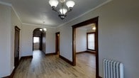 This single-floor space showcases a blend of classic architectural character and timeless design. The interior is defined by rich, dark wood trim that frames doorways and baseboards throughout, creating a striking contrast against the light gray walls. Crown molding runs along the ceilings, adding an elegant finishing detail to each room.

The main hallway serves as the central artery of the space, featuring hardwood flooring that extends seamlessly into the adjoining rooms. A decorative chandelier hangs from the ceiling, providing both illumination and a classic aesthetic focal point. An arched doorway at the end of the hallway adds architectural charm and a sense of depth, leading further into the space.

A generously sized adjacent room is visible through a wide, dark-trimmed entryway. This room benefits from natural light streaming through a window, and is also fitted with a ceiling fan for climate comfort. The hardwood floors continue here, maintaining a cohesive look throughout.

A cast iron radiator is visible along one wall, a nod to the building's historical character. Overall, the space carries a warm, traditional ambiance underscored by its period-appropriate architectural details, making it both functional and full of timeless charm.