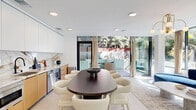 Located on Ohio Avenue in Los Angeles's desirable West LA neighborhood, this thoughtfully designed single-floor space blends contemporary architecture with refined interior finishes. The open-plan layout seamlessly connects the kitchen, dining, and living areas, creating a bright and cohesive environment flooded with natural light from expansive floor-to-ceiling windows and glass sliding doors.

The kitchen is a standout feature, boasting a dramatic full-length marble backsplash with bold veining, warm natural wood lower cabinetry, sleek white upper cabinets, and stainless steel appliances. A large oval dining table with upholstered cream chairs sits centrally, offering an inviting gathering space directly adjacent to the kitchen.

The living area is anchored by a sculptural curved blue velvet sofa positioned before panoramic windows, creating a light-filled lounge atmosphere with views of surrounding greenery. A distinctive glass-block partition wall adds a retro-modern architectural detail, diffusing soft light while defining the space.

Terrazzo flooring runs throughout the interior, contributing a polished, timeless aesthetic. The space opens directly onto a private patio featuring a raised concrete garden planter filled with ornamental grasses and plants, framed by mature trees. The patio includes casual seating and is enclosed by walls and a gated entry, offering a tranquil outdoor retreat.