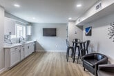 Located in Memphis, TN, this single-level community room offers a clean, contemporary design ideal for gatherings and shared use. The open layout is finished with light hardwood flooring throughout, neutral gray walls, and recessed lighting that keeps the space bright and welcoming.

The kitchenette is a standout feature, showcasing white shaker cabinetry with matte black hardware, striking marble-veined countertops and a matching backsplash, a stainless steel undermount sink with a black matte faucet, and a window overlooking neighboring buildings. A dishwasher is integrated into the cabinetry, adding everyday functionality.

The main gathering area provides flexible seating arrangements, including black leather lounge chairs and a loveseat, as well as a high-top round table with industrial-style metal bar stools. A wall-mounted flat-screen television anchors one wall, making the space well-suited for presentations or casual viewing. Contemporary artwork adds pops of color against the neutral palette.

The room's open floor plan allows for versatile use, from social events to meetings. A separate door provides access to an additional area, enhancing the room's adaptability. The overall ambiance is modern, clean, and inviting.