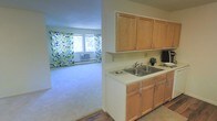 This single-floor unit features a practical, open layout that connects the kitchen and living area seamlessly. The kitchen is outfitted with light wood cabinetry offering ample upper and lower storage, a double-basin stainless steel sink, and light-toned countertops. The hardwood-style flooring in the kitchen transitions into plush carpeting in the adjacent living area, creating a clear yet open delineation between the two spaces. The living area is bright, benefiting from a large window dressed with colorful patterned curtains that allow natural light to fill the room. A wall-mounted air conditioning unit and baseboard heating provide year-round climate control. The neutral wall tones throughout the space contribute to a clean, understated atmosphere, offering a versatile backdrop for a variety of interior styles. Overall, the layout is efficient and functional, making good use of the available space while maintaining an open, airy feel between the kitchen and living zones.