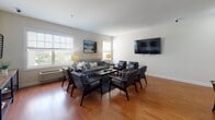 Located at 255 E Grand Ave in Rahway, New Jersey, this single-floor communal space offers a thoughtfully designed layout that balances comfort and functionality. The modern kitchen is outfitted with sleek, dark flat-front cabinetry, crisp white countertops, a textured stone tile backsplash, stainless steel appliances, and warm hardwood-style flooring — a combination that gives the space a contemporary, polished feel. The open lounge area is bright and inviting, featuring large windows that allow generous natural light to fill the room. Comfortable seating arrangements, including a sofa and mid-century modern accent chairs, are centered around a coffee table, creating an ideal setting for relaxation or casual gatherings. A wall-mounted flat-screen television adds a practical amenity to the space. The dining area accommodates multiple tables with industrial-style metal chairs, complemented by tasteful artwork and another wall-mounted display screen, making it well-suited for group meals or meetings. The bathroom is clean and simply appointed, with a white vanity, a tub-shower combination with curtain, and hardwood-style flooring that ties in with the rest of the space. Throughout, neutral tones and warm wood floors create a cohesive, welcoming atmosphere.