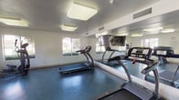 Located at 18005 W Anne's Cir in Canyon Country, CA, this single-floor fitness center offers a well-equipped workout facility designed to support a range of exercise routines. The space is organized into distinct activity zones, providing ample room for both cardio and strength training without feeling congested.

The cardio area is home to multiple treadmills, an elliptical trainer, and a recumbent stationary bike, all positioned to take advantage of the natural light streaming in through large windows. These windows also frame views of a neighboring outdoor pool and landscaped grounds, creating an inviting atmosphere during workouts. A wall-mounted television is available for entertainment.

The strength training section features a multi-station cable weight machine, offering a variety of upper and lower body exercise options. Full-length mirrors line the walls throughout the facility, enhancing the sense of openness and allowing users to monitor their form during exercise.

The flooring throughout is a durable, speckled blue rubberized surface well-suited to a high-activity environment. Overhead flush-mount lighting ensures the space is consistently and evenly illuminated. Restroom facilities are accessible directly from within the fitness center. Fitness center rules and guidelines are posted near the entrance, and the facility maintains a no-smoking policy. Overall, the space is clean, functional, and thoughtfully arranged for community fitness use.