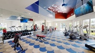 Located at 700 Forest Park Boulevard in Oxnard, California, this fitness center is a single-floor exercise facility offering a generous and open workout environment. The space makes an immediate visual impact with its striking blue and white scalloped-pattern carpet, bold red and blue color-blocked ceiling, and motivational typographic lettering overhead — all contributing to an energetic and dynamic atmosphere.

The facility is comprehensively equipped for a full-body workout experience. A dedicated strength training zone features a variety of selectorized resistance machines with red upholstery, a dumbbell rack, and free weight benches. The cardio zone, positioned near floor-to-ceiling windows, houses treadmills, ellipticals, stationary bikes, and a rowing machine, all of which benefit from abundant natural light and views of the surrounding landscaping and palm trees.

Full-length mirrors line the walls, visually expanding the space and supporting proper form during exercise. A ceiling-mounted flat-screen television and pendant lighting add to the amenities. A wall-mounted rack near the entry displays yoga mats and exercise balls, and glass-paned double doors connect the fitness floor to an adjacent lounge or community room. Recessed lighting throughout ensures a bright, welcoming interior at all hours.