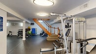 Located at 855 King Arthur Dr in Fayetteville, NC, this dedicated exercise room delivers a comprehensive fitness environment across a single floor. The space features durable dark rubber flooring throughout, providing both safety and functionality for a wide range of workout activities. The interior is finished with clean white walls and flush-mount ceiling light fixtures that ensure bright, even illumination across the entire area.

The layout is thoughtfully organized to accommodate multiple workout zones. A multi-station cable weight machine takes center stage alongside an adjustable bench, supporting strength training exercises. Complementing this is a full rack of dumbbells, a barbell with a power rack, and several stability and medicine balls, catering to both free-weight and functional training routines. The cardio section features multiple stationary bikes and elliptical machines, offering ample options for cardiovascular workouts.

A large wall mirror in one section expands the sense of space and allows users to monitor their form during exercise. A staircase with warm wood banisters connects the gym to the floor above, and a private door provides convenient access. A wall-mounted television and a posted information board round out the amenities, making this exercise room a fully self-contained and practical fitness facility.