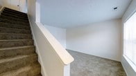 This two-story space features a straightforward and functional layout connected by a carpeted staircase with a clean white banister and newel post. The stairs are finished with a tan, textured carpet that continues onto the upper level, providing a cohesive and comfortable feel underfoot. The upper landing or hallway area is open and simply appointed, with smooth white walls and a lightly textured ceiling that contributes to a clean, neutral aesthetic. The flooring on the upper level is finished in the same understated carpet, creating a consistent look throughout. The walls are painted in a soft, neutral white tone, lending the space a bright and airy quality. The open railing design along the staircase adds a sense of openness between the two floors. Overall, the space presents a simple, well-maintained interior with a clean palette, making it a versatile foundation that can be easily personalized or furnished to suit a variety of styles and needs.