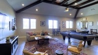 This generously sized single-floor space functions as a combined game room and lounge, offering an inviting layout suited for both recreation and relaxation. The most striking architectural feature is the vaulted ceiling adorned with dark-stained exposed wood beams, which add rustic character and a sense of vertical openness to the room. Recessed lighting and a pendant ceiling fan provide both functional illumination and aesthetic detail overhead.

The space is lined with multiple windows along three walls, allowing abundant natural light to fill the interior and creating a bright, airy atmosphere throughout the day. Neutral-toned walls complement the warm tones of the tile flooring, which is partially covered by a large, richly patterned area rug that anchors the seating area.

The lounge zone features a tan upholstered sofa accented with decorative pillows, accompanied by leather armchairs and a wooden coffee table. Framed artwork on the walls adds a curated, finished look. On the opposite side of the room, a full-sized pool table serves as the centerpiece of the game area, with a cue rack nearby. Bar-height seating near the windows provides an additional casual gathering spot, rounding out the room's versatile appeal.