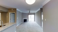 This single-floor residence offers a thoughtfully designed layout with a welcoming, open-concept living area at its heart. The spacious living room is finished with plush wall-to-wall carpeting and neutral gray walls accented by crisp white crown molding, creating a clean and polished aesthetic. A classic fireplace serves as a central focal point, adding warmth and character to the space. A ceiling fan overhead provides both function and style, while a sliding glass door framed by vertical blinds allows natural light to pour in and offers a direct connection to an outdoor space beyond. The flush mount ceiling light adds a modern, understated touch. Adjacent to the living area, the kitchen is distinguished by its granite countertop surfaces, hinting at quality finishes throughout. The open sightlines between the kitchen and living areas enhance the sense of spaciousness and make the layout ideal for everyday living and entertaining. The unit is contained on a single level, ensuring convenient and accessible flow throughout. Overall, the space presents a comfortable and functional living environment with a balance of practical design and appealing interior finishes.