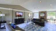 This single-floor space showcases a thoughtfully designed living room that combines comfort with modern style. The room's most striking architectural feature is its vaulted ceiling, which adds a sense of height and openness to the space. Warm gray walls provide a neutral backdrop that complements the room's carefully chosen furnishings and accents.

The living area is anchored by a pair of dark charcoal sofas adorned with blue and green accent pillows, arranged around a geometric glass-and-chrome coffee table. A bold blue and white chevron-patterned area rug defines the seating area and adds a dynamic visual element to the space. A wall-mounted flat-screen television sits atop a black media console, positioned across from the seating arrangement for optimal viewing.

A standout feature is the natural stone fireplace, which rises along one wall and adds a rustic yet refined character to the room. Recessed lighting in the ceiling, combined with a window that allows natural light to filter in, ensures the space feels bright and inviting. A decorative round mirror and a small bar-height table near the window add finishing touches that enhance the room's overall charm.