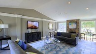 This single-floor space showcases a thoughtfully designed living room that combines comfort with modern style. The room's most striking architectural feature is its vaulted ceiling, which adds a sense of height and openness to the space. Warm gray walls provide a neutral backdrop that complements the room's carefully chosen furnishings and accents.

The living area is anchored by a pair of dark charcoal sofas adorned with blue and green accent pillows, arranged around a geometric glass-and-chrome coffee table. A bold blue and white chevron-patterned area rug defines the seating area and adds a dynamic visual element to the space. A wall-mounted flat-screen television sits atop a black media console, positioned across from the seating arrangement for optimal viewing.

A standout feature is the natural stone fireplace, which rises along one wall and adds a rustic yet refined character to the room. Recessed lighting in the ceiling, combined with a window that allows natural light to filter in, ensures the space feels bright and inviting. A decorative round mirror and a small bar-height table near the window add finishing touches that enhance the room's overall charm.