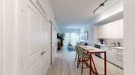 This thoughtfully designed one-bedroom, one-bathroom unit offers an efficient and comfortable single-floor layout. Light wood-style flooring flows throughout, lending a clean, cohesive look to the entire space. The open-concept kitchen features floor-to-ceiling white cabinetry, stainless steel appliances including a French door refrigerator, a range, microwave, and dishwasher, complemented by track lighting overhead. A compact bar-height dining table with two chairs serves as a casual dining and gathering area adjacent to the kitchen.

The living room is bright and inviting, benefiting from natural light streaming through a sliding glass door that opens to an outdoor patio area. White walls and light finishes keep the space feeling airy and open. A sliding barn-style door adds a modern architectural touch near the entryway, where two closets provide practical storage.

The bedroom is a calm retreat with a window allowing natural light, neutral walls, and carpet-style area rugs softening the flooring. The bathroom features a full soaking tub with a tiled surround in a neutral beige palette, a curved shower rod, and clean white fixtures. A hallway connects the main living areas to the private bedroom and bathroom, maintaining a functional and organized flow throughout the unit.