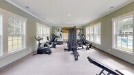Located at 102 Lusk Lane in Pooler, Georgia, this fitness center offers a well-appointed workout facility spread across two dedicated exercise rooms on a single floor. The space is finished with soft sage-green walls, crisp white crown molding, and neutral carpeted flooring, creating a clean and inviting atmosphere throughout.

The main exercise room is generously sized and filled with an impressive array of equipment, including elliptical trainers, stationary bikes, a treadmill, a multi-station cable machine, a rowing machine, and adjustable weight benches. Multiple large white-framed windows line the walls, flooding the space with abundant natural light and offering views of the surrounding outdoor area, including glimpses of a pool. Recessed ceiling lighting complements the natural illumination, ensuring a bright environment at any time of day.

The second exercise room provides a more intimate setting for strength and flexibility training, featuring a dumbbell rack with a full set of free weights and an adjustable workout bench. A glass-paneled exterior door offers direct outdoor access and additional light. A built-in bench and cubby area with wainscoting provides a convenient space to store belongings. Colorful artwork adorns the walls of both rooms, adding a welcoming and motivating touch to the overall environment.