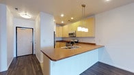 This single-floor space centers around a well-appointed open kitchen that seamlessly integrates with the surrounding living area. The kitchen is anchored by a peninsula with a warm brown countertop that doubles as a breakfast bar, housing a stainless steel double sink and offering generous prep space. A trio of pendant lights hangs above the peninsula, providing both task lighting and a stylish focal point that draws the eye upward to the white ceiling with recessed lighting.

Light natural wood cabinetry lines the kitchen walls, offering ample upper and lower storage. Stainless steel appliances, including a gas range, built-in microwave, and refrigerator, complement the cabinetry with a modern, cohesive aesthetic. The countertops throughout carry a consistent warm brown tone that ties the space together.

Dark hardwood flooring extends throughout, adding depth and contrast to the bright white walls and lighter cabinetry. The open layout promotes a sense of flow and connectivity, with a visible entryway corridor adding to the functional design. Recessed ceiling lights ensure the space remains well-lit throughout.