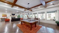 Located at 7035 Pickwell Dr in San Antonio, Texas, this single-story space makes a striking impression through its bold design choices and generous open layout. The kitchen is a standout, featuring crisp white upper cabinetry contrasted by dark lower cabinets, gleaming granite countertops, and a vivid red subway tile backsplash that adds dramatic flair. A large island with seating for several, topped with matching granite and illuminated by industrial-style pendant lights, anchors the space. Stainless steel appliances complete the well-appointed kitchen. The expansive living area is anchored by a stone fireplace with a reclaimed wood mantle and a wall-mounted television, flanked by large glass doors that invite natural light and outdoor views. Exposed wooden ceiling beams and a tray ceiling overhead lend architectural character, while a statement chandelier adds an artistic focal point. Bold red leather seating and a dark leather sofa create a lively, layered atmosphere throughout. A game area features a pool table beneath a sliding barn door accent wall. A dedicated workspace area offers individual desk stations separated by colorful partitioned panels in red and yellow. Wood-look tile flooring flows throughout, tying together the space's energetic and eclectic personality.