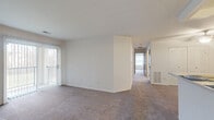 This single-floor residence offers a clean, functional layout designed for comfortable everyday living. The open living area is finished with plush wall-to-wall carpeting and crisp white walls, creating a bright and neutral backdrop. A sliding glass door fills the space with natural light and provides direct access to an outdoor balcony or patio, with vertical blinds offering privacy when needed. A ceiling fan overhead aids in air circulation throughout the living space.

The kitchen is positioned adjacent to the living area and features a pass-through breakfast bar that connects the two spaces. It is equipped with white cabinetry, a gas range, and a full-size refrigerator, with hard surface flooring that contrasts the carpeted living areas.

A hallway leads to the bedrooms, each offering generous floor space, carpeted floors, and ample closet storage — including what appears to be a walk-in closet in at least one room. The primary bedroom includes an en-suite bathroom with a vanity, mirror, and shower with curtain.

An additional full bathroom is accessible from the hall, also featuring hard surface flooring. Throughout, the space maintains a consistent neutral palette with white walls and warm-toned carpet, offering a versatile canvas ready for personalization.