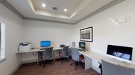 This single-story business center in Katy, Texas offers a well-appointed, professionally designed environment suited for productivity and comfort. The space opens into a grand lounge area distinguished by soaring ceilings, polished concrete floors, and an elegant chandelier that anchors the room. Striped upholstered seating arranged around a marble-top coffee table creates an inviting gathering area, while recessed lighting and clerestory windows bathe the space in natural light.

Adjacent to the lounge, a dedicated computer room provides multiple workstations along continuous granite-topped desks, each equipped with desktop computers and ergonomic task chairs featuring orange accent cushions. A tray ceiling with recessed lighting adds architectural interest to this workspace, which also includes access to a printer. Natural light filters in through slatted blinds on the windows, complementing the rich wood-look flooring.

A secondary, more intimate workstation area offers a quieter setting with a single iMac workstation, accessible through glass-paneled doors that maintain visual connectivity to the lounge. Throughout the space, neutral wall tones, framed artwork, and consistent design elements create a cohesive, polished aesthetic that balances functionality with a welcoming, upscale ambiance.