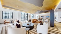 Located at 45 Northeast 27th Street in Miami's vibrant Edgewater neighborhood, this thoughtfully designed amenity space spans a generous single floor, offering a refined setting for socializing and relaxation. The interior is immediately distinguished by its striking ceiling treatment — a sweeping, cloud-like mural rendered in gold, gray, and blue tones that serves as an artistic focal point throughout the space.

The open-plan layout encompasses two interconnected zones. The main lounge area features an array of plush seating, including cream boucle armchairs and a rich cognac leather sectional sofa, all arranged atop layered geometric-patterned area rugs that complement the terrazzo-tiled flooring. Curated abstract artwork adorns the walls, contributing to a gallery-like ambiance.

Floor-to-ceiling glass walls and steel-framed windows flood the interior with abundant natural light, while offering views of an adjacent outdoor terrace and the surrounding Miami cityscape. Ambient cove lighting further enhances the warm atmosphere, particularly in the evening.

A secondary lounge and media area features wood-paneled walls, a wall-mounted television, and casual seating arrangements, providing a more intimate gathering space. The overall design balances contemporary elegance with artistic expression, creating an inviting and visually dynamic environment.