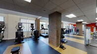 This fitness center offers a comprehensive workout environment spread across a single, open floor plan. The space is defined by its distinctive multi-toned rubber flooring — a combination of dark grey, black, and gold tiling — which delineates different functional zones throughout the facility. Structural brick columns punctuate the interior, adding architectural character while dividing the space into clearly organized workout areas.

The cardio section is well-stocked with a row of treadmills equipped with individual monitors, alongside elliptical trainers and stationary bikes, all positioned near large windows fitted with horizontal blinds that allow ample natural light to fill the space. A full-length mirror along one wall visually expands the area and supports form-checking during exercise.

The strength training zone features a multi-station cable machine, a Smith machine, adjustable benches, and a selection of Life Fitness selectorized equipment. A dedicated functional training area includes stability balls, medicine balls, plyo boxes, and additional free-weight accessories. Bold orange accent walls provide vibrant visual contrast against the neutral tones of the ceiling and walls.

The facility is accessed through a glass-paneled entry with black framing, offering a clean and professional aesthetic. Recessed panel lighting throughout ensures consistent, even illumination across all workout zones, creating a practical and motivating training environment.
