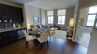 This well-appointed single-floor residence showcases a thoughtfully designed open-concept layout that seamlessly integrates the living, dining, and kitchen areas into one cohesive space. Rich, dark hardwood floors run throughout, providing a warm and elegant foundation that contrasts beautifully with the neutral-toned walls.

The dining area features a round dark-wood table paired with crisp white upholstered chairs, creating an inviting setting for meals and gatherings. Just beyond, the living area is anchored by a comfortable sofa accented with yellow and grey throw pillows, complemented by a geometric patterned area rug in gold and white tones that adds visual interest and warmth underfoot.

Large windows with roller shades allow generous natural light to flood the space while offering views of the surrounding cityscape. The bright, airy atmosphere is further enhanced by well-placed floor and table lamps that create a warm, layered lighting scheme throughout.

A glimpse of the kitchen reveals modern cabinetry and stainless steel appliances, hinting at a functional and stylish culinary space. The overall ambiance of this residence is modern yet comfortable, with a cohesive neutral and gold color palette that lends a sophisticated, contemporary feel to every corner.