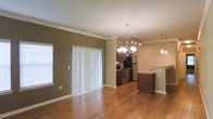 This single-floor residence offers a thoughtfully designed open-concept layout that seamlessly connects the living area to the kitchen. Warm hardwood flooring extends throughout the main living space, complemented by neutral olive-toned walls and crisp white crown molding that adds a polished architectural detail to each room.

The living area is spacious and inviting, featuring a large sliding glass door dressed with vertical blinds that allows natural light to filter in while providing access to the outdoors. A side window further brightens the space, contributing to an airy and comfortable atmosphere.

The kitchen is a standout feature of this home, outfitted with rich dark wood cabinetry, granite countertops, and a full suite of stainless steel appliances including a refrigerator, stove, and microwave. A breakfast bar peninsula provides a casual dining option and serves as a natural divider between the kitchen and living areas. Overhead, an elegant chandelier anchors the dining space, while track lighting and a coordinating pendant light illuminate the kitchen with both style and function.

The overall ambiance of this residence is warm and modern, blending classic architectural touches with contemporary finishes to create a comfortable and visually appealing living environment.