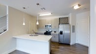 This well-appointed two-story space showcases a thoughtfully designed interior with modern finishes and a clean, contemporary aesthetic. The open-concept kitchen serves as a central highlight, featuring stainless steel appliances including a gas range, microwave, and a French door refrigerator with an in-door water and ice dispenser. Flat-panel cabinetry in a warm taupe finish lines the walls, complemented by a subtle mosaic tile backsplash that adds texture and visual interest. A built-in wine rack above the refrigerator offers a practical yet stylish storage solution for entertaining enthusiasts. The expansive white countertop island provides generous workspace and doubles as a casual dining or gathering spot, illuminated overhead by sleek pendant lights that hang at varying heights for a modern touch. Rich, dark hardwood-style flooring runs throughout, grounding the space and contrasting beautifully with the light-toned cabinetry and walls. The layout is open and airy, with clean sightlines that enhance the sense of space. Recessed and flush-mount lighting ensures the kitchen is well-lit throughout. Spanning two floors, this space offers a versatile and functional layout suited to a variety of lifestyles.