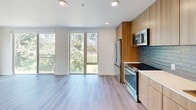 Located at 3601 S Alaska St in Seattle's vibrant Columbia City neighborhood, this thoughtfully designed 3-bedroom, 2-bathroom unit offers a clean, modern aesthetic throughout its well-organized single-floor layout. Light hardwood-style flooring runs continuously through the space, complementing the bright white walls and creating a cohesive, contemporary feel.

The open-concept kitchen and living area serves as the heart of the unit, featuring floor-to-ceiling windows and a glass sliding door that invite abundant natural light and frame lush green outdoor views. The kitchen is outfitted with warm wood cabinetry, a gray subway tile backsplash, white quartz countertops, and a full suite of stainless steel appliances including a range, microwave, refrigerator, and dishwasher.

The primary bedroom includes a stylish sliding barn door leading to a dedicated closet. Additional bedrooms offer generous storage with double sliding-door closets and large windows overlooking greenery. Two bathrooms serve the unit — one featuring a soaking tub with full tile surround, and the other with a modern wood-grain vanity, undermount sink, and lighted mirror. An in-unit washer is also present, adding everyday convenience to this well-appointed Seattle home.