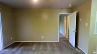 This single-story space features a straightforward layout with carpeted flooring throughout, providing a warm and understated aesthetic. The room pictured is finished with warm tan-painted walls and white trim, creating a clean contrast along the baseboards and door frames. Overhead lighting is provided via a ceiling-mounted fixture, illuminating the space adequately. A standard electrical outlet is visible along the lower portion of the wall, reflecting the room's practical design.

An open doorway leads directly into a hallway or adjoining area, where additional rooms are accessible. Through this opening, a second doorway is visible, suggesting a connected layout with multiple rooms branching off a central corridor. The walls in the adjoining area are painted in a cooler gray-blue tone, offering a subtle visual distinction between spaces.

The carpeting throughout appears to be a medium-pile, neutral-toned material consistent across the visible rooms, contributing to a cohesive interior finish. The overall ambiance is functional and move-in ready, with a clean, no-frills design suited for comfortable everyday living. The space presents a blank canvas, well-suited for personalization through furnishings and décor.
