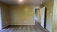This single-story space features a straightforward layout with carpeted flooring throughout, providing a warm and understated aesthetic. The room pictured is finished with warm tan-painted walls and white trim, creating a clean contrast along the baseboards and door frames. Overhead lighting is provided via a ceiling-mounted fixture, illuminating the space adequately. A standard electrical outlet is visible along the lower portion of the wall, reflecting the room's practical design.

An open doorway leads directly into a hallway or adjoining area, where additional rooms are accessible. Through this opening, a second doorway is visible, suggesting a connected layout with multiple rooms branching off a central corridor. The walls in the adjoining area are painted in a cooler gray-blue tone, offering a subtle visual distinction between spaces.

The carpeting throughout appears to be a medium-pile, neutral-toned material consistent across the visible rooms, contributing to a cohesive interior finish. The overall ambiance is functional and move-in ready, with a clean, no-frills design suited for comfortable everyday living. The space presents a blank canvas, well-suited for personalization through furnishings and décor.