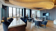 This elevated amenity floor presents a carefully curated collection of social and leisure spaces united by a cohesive, upscale design language. The grand lobby lounge serves as the centerpiece, anchored by a towering dark metallic feature wall that extends to the ceiling and is flanked by sculptural, multi-arm pendant chandeliers. Symmetrically arranged high-back chairs in deep burgundy and white sit atop a boldly patterned area rug, while additional seating groups line the perimeter beneath vibrant large-scale artwork. Light hardwood flooring runs throughout, providing a warm contrast to the dramatic dark accents.

A hallway corridor with dark accent walls, curated wall-mounted panels, and steel-framed glass doors leads to additional amenity spaces. The social lounge and dining area features a sculptural wave-form ceiling installation in warm gold tones, a marble-topped kitchen bar, round dining tables with navy chairs, and a billiards table. A polished metallic sculpture adds an artistic focal point, while floor-to-ceiling windows flood the space with natural light and panoramic city views. A dedicated lounge nook offers deep leather sectional seating positioned directly before expansive windows, framing the urban skyline beyond.