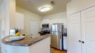 Located at 935 Stone Lion Dr in Durham, NC, this single-floor unit offers a well-organized layout with distinct living, kitchen, bedroom, and bathroom areas. The kitchen is a functional galley-style space outfitted with white shaker cabinetry extending to the ceiling, dark granite countertops, and a full suite of stainless steel appliances including a refrigerator, electric range, and over-the-range microwave. A peninsula counter provides additional workspace and creates a natural division between the kitchen and the adjacent living area. Hardwood-style flooring runs throughout the kitchen and bathroom, adding warmth and continuity. The living area features plush carpeting, crown molding, a ceiling fan with light fixture, and a window that allows natural light to fill the space. The bedroom mirrors this comfort with carpeted flooring, a ceiling fan, and a window offering outdoor views. The bathroom is appointed with a wide, dark-framed mirror, granite-topped white vanity with an undermount sink, and a shower-tub combination with a curtain rod. Throughout the unit, neutral wall tones and white trim create a clean, cohesive aesthetic.