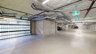This large-scale underground parking garage offers a generous, well-organized layout across a single basement level. The floor plan is elongated and open, accommodating numerous clearly marked vehicle stalls throughout the space. The structure is built with exposed concrete columns and walls, giving the facility a solid, utilitarian character. Overhead, an extensive network of mechanical pipes, conduits, and sprinkler systems runs along the spray-foam-insulated ceiling, with linear fluorescent lighting providing consistent illumination throughout.

Access to the garage is controlled via a robust metal security gate, ensuring a secure entry and exit point. The smooth, light-toned concrete floor is clearly marked with painted stall designations and directional indicators. Electric vehicle charging stations are installed at select stalls, reflecting a modern infrastructure upgrade. A dedicated, chain-link-enclosed bicycle storage area is conveniently situated within the garage, offering secure parking for cyclists. Additional utility areas, including electrical panels and storage rooms, are integrated along the perimeter walls. Emergency exit signage and safety equipment, including fire suppression components, are prominently visible throughout, underscoring the facility's commitment to safety and code compliance.