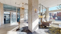 This refined building lobby makes an immediate impression with its expansive travertine stone walls and matching stone flooring, lending the space a timeless, upscale character. The single-floor layout is thoughtfully organized into distinct functional zones that flow seamlessly from one to the next. At the entrance, a set of glass-and-aluminum doors opens into a grand reception corridor framed by monumental stone columns and stanchion barriers, conveying a sense of curated arrival. Gold-trimmed elevator doors with brass call panels line one side of the corridor, their warm metallic finish complementing the travertine surroundings and a series of elegant brass chandeliers and wall sconces that illuminate the space throughout. Adjacent to the elevator bank, rich wood-paneled walls add warmth and contrast to the otherwise light stone palette. The lounge area serves as a welcoming social hub, featuring a curated arrangement of contemporary sofas, armchairs, and accent tables atop a patterned area rug with grey, blue, and gold tones. Expansive floor-to-ceiling windows flood this seating area with abundant natural light and offer clear views of the surrounding streetscape and mature trees. The overall ambiance is polished and professional, balancing classic architectural materials with modern design sensibilities.